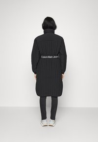 Abrigo largo acolchado negro con cuello alto y cinturón elástico con el logo de Calvin Klein Jeans. Llevado sobre pantalones ajustados grises y calzado ligero.