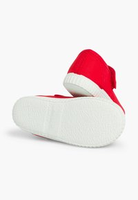 Pantofi din pânză roșie cu tălpi din cauciuc alb, având un design simplu și o curea cu arici. Textura pare netedă, cu o talpă modelată.
