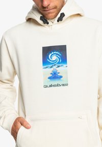 Krém színű kapucnis pulóver, elülső zsebbel, kék és fekete grafikai mintázattal, amely örvénylő vizet és hegyeket ábrázol. A "Quiksilver" márkanevének felirata alatta található.