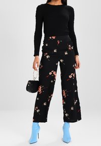 Pantalons noirs à imprimé floral avec jambes larges, présentant un motif floral coloré, associés à un haut noir ajusté et des chaussures à talons hauts bleues.
