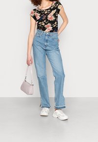 Haut noir à motif floral avec manches courtes, jeans taille haute bleu clair, baskets blanches et petit sac à main rose. Modélise une tenue décontractée.