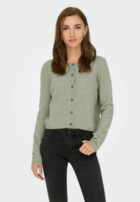 Zaļa kardigāna veste, kas izgatavota no mīksta adījuma materiāla, ar apaļu kakla izgriezumu, priekšējo pogu aizdari un garām piedurknēm, apvienota ar melniem džinsiem.