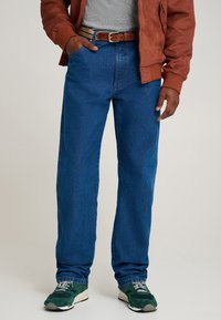 Blugi de denim albastru cu croială dreaptă, buzunare și ușoară decolorare. Asortați cu un tricou gri, o jachetă bomber maro și adidași verzi.