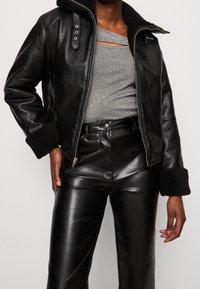 Giacca in pelle nera con cappuccio, polsini foderati in pelliccia e zip metallica. Indossata sopra una maglia grigia, abbinata a pantaloni neri in finta pelle lucida.