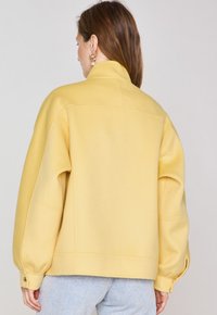 Yellow, oversized jacket with a high collar, dropped shoulders, and buttoned cuffs. Smooth fabric with minimal stitching detail.