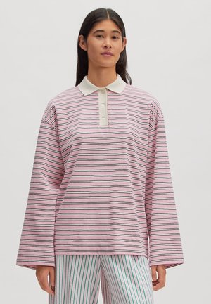 Woman wearing a long-sleeve pink polo shirt with thin black stripes and white collar, paired with white pants with green vertical stripes.