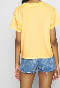 Nainen, jolla on kirkkaan oranssi lyhythihainen paita ja siniset farkkushortsit, joissa on reunat, seisoo selkä kameraan päin.