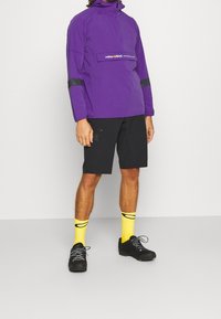 Giacca a pullover viola con zip a metà e dettagli neri, abbinata a pantaloni corti neri, calzini gialli con strisce nere e scarpe nere.