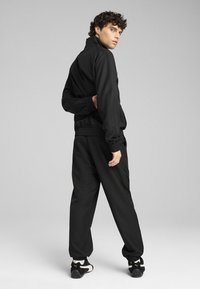 Black athletic tracksuit with a zip-up jacket and elastic cuffs, paired with loose-fitting pants. Displayed on a model from the back.