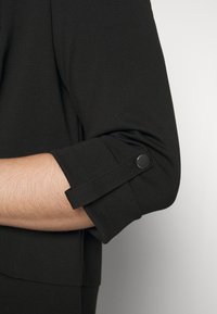 Top noir ajusté avec des manches roulées et un détail de bouton. Texture lisse et design simple sans motifs ni embellissements.
