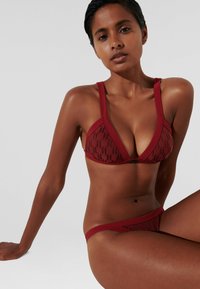 Raudonas bikini komplektas, kurį sudaro trikampis liemenėlė su raštuotu audiniu ir plačiomis petnešėlėmis, suderintas su atitinkamomis žemomis iškirptėmis kelnaitėmis. Glotni tekstūra.