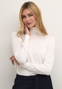 Pulover alb cu guler înalt, realizat dintr-un material moale, cu mâneci lungi și croială mulată, asortat cu pantaloni de culoare bleumarin închis.