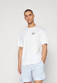 Hvit bomull T-skjorte med korte ermer, med en liten Reebok-logo i svart og rødt på brystet. Båret med lyseblå perforerte shorts.