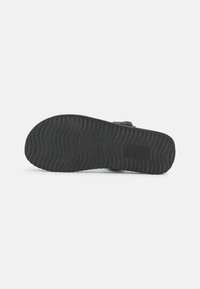 Black shoe sole with a wavy texture and grooved pattern, made from rubber. Features a rounded toe and flat profile.