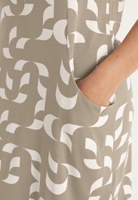 Grauer Stoff mit glatter Textur, der ein markantes weißes abstraktes Muster aufweist. Enthält eine seitliche Tasche, die das praktische Design unterstreicht.