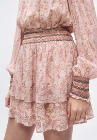 Robe longue à manches longues en tissu transparent avec des motifs floraux complexes roses et bruns, taille et poignets froncés, jupe à volants superposés, main reposant le long du corps.