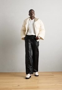 Krémová péřová bunda, bílá košile na knoflíky, černé široké kalhoty z umělé kůže a bílé loafers s černými podrážkami; minimalistický design.