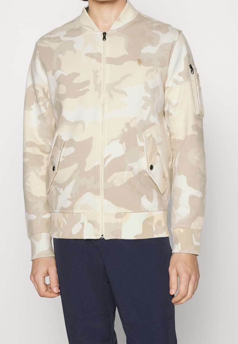 Giacca mimetica in tonalità crema, con zip frontale, collo alla coreana, due tasche laterali e una tasca sulla manica. Tessuto morbido e liscio al tatto.