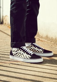 Baskets à motif damier noir et blanc, avec des lacets blancs et une semelle blanche, associées à un pantalon noir retroussé sur une surface en béton.