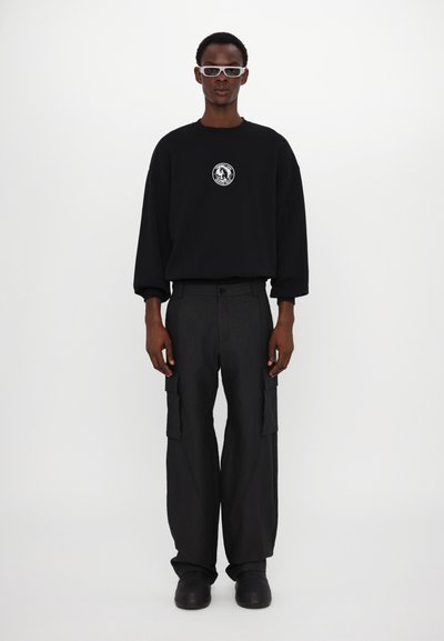 Sweatshirt noir oversize avec un graphique circulaire sur la poitrine, accompagné d'un pantalon cargo noir avec poches latérales et de chaussures noires à enfiler.