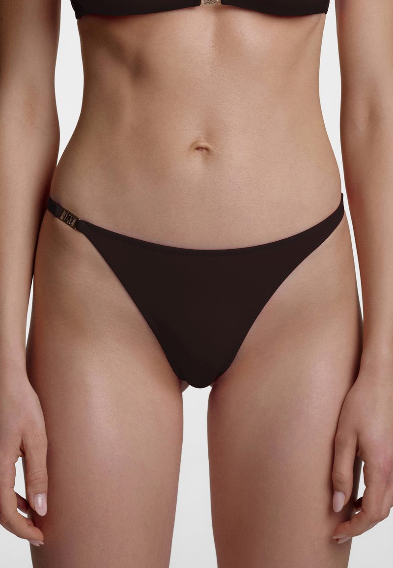 Partie médiane d’une femme portant un bas de bikini noir avec un détail de fermoir métallique sur le côté gauche sur un fond uni.