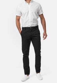 Pantaloni in cotone neri con vestibilità slim, dotati di bottone in vita, tasche laterali e una texture liscia, abbinati a una camicia bianca a maniche corte.