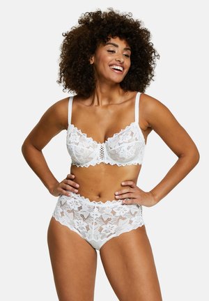 Donna sorridente con capelli ricci che indossa un reggiseno in pizzo bianco e mutandine in pizzo a vita alta abbinate, con le mani appoggiate sui fianchi.