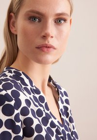 Blusa bianca con grandi pois blu navy, con scollatura a V e maniche corte. Tessuto liscio con una leggera lucentezza.