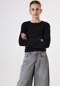 Schwarzes Langarmshirt mit verstreuten Strasssteinen, kombiniert mit hochgeschnittenen, locker sitzenden grauen Jeans. Glatte Textur und taillierte Passform.