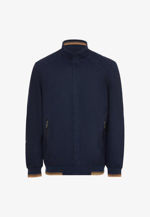 Námořnická modrá bunda vyrobená z látky s vysokým límcem, dvěma bočními kapsami na zip, žebrovanými manžetami a kontrastním zlatohnědým akcentem na spodním lemu.