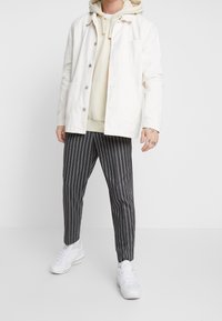 Valkoinen kangastakki beige hupparin päällä, yhdistettynä tummiin raidallisiin housuihin ja valkoisiin lenkkareihin. Sisältää napillisen sulkemisen ja taskut.