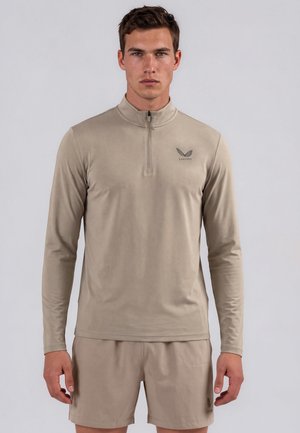 Homme portant une chemise beige à manches longues zippée et un short assorti, debout devant un fond gris uni.