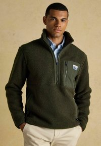 Fleece pullover in dark green with textured fabric, zippered chest pocket, stand collar, and side pockets, worn over a light blue shirt.