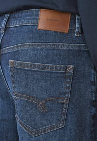 Ciemnogranatowe jeansy z denimu mają skórzaną naszywkę z napisem „DENIM SUPPLY” oraz kontrastujące pomarańczowe przeszycia na tylniej kieszeni.