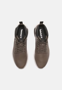 Hnědé kožené tenisky s texturovanými akcenty, kulatá špička, kontrastní bílá podrážka a černý interiér. Tkaničky jsou zajištěny pomocí průvleků.