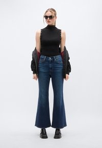 Μαύρο κορμάκι χωρίς μανίκια με κουμπιά κλιπ στους ώμους, μπλε τζιν καμπάνα και μαύρα χοντρά μποτάκια. Το μοντέλο φοράει σκούρα γυαλιά ηλίου.