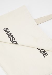 Gros plan d'un sac fourre-tout en toile beige plié avec texte imprimé noir partiellement visible, montrant un tissu solide et une poignée.