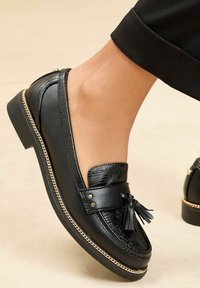 Mustat loafers, joissa on teksturoitu etuosa, tupsuyksityiskohta, kultasävyinen ketjuviimeistely ja matala korko. Huomattavissa sileä nahka ja kiiltävät yksityiskohdat.