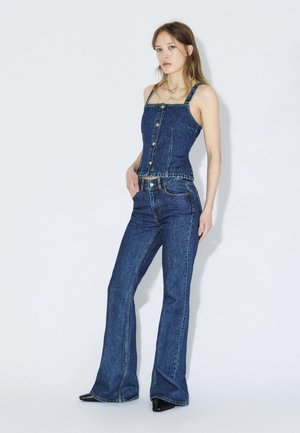 Vrouw staat tegen een witte muur, draagt een denim korsettetop met knopen en hooggetailleerde uitlopende jeans met zwarte puntige schoenen.