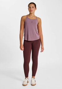 Débardeur sportif violet clair à fines bretelles, associé à des leggings bordeaux et des baskets blanches. Le débardeur arbore un détail logo subtil.