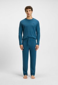Jeune homme debout pieds nus portant un haut à manches longues bleu assorti et un pantalon à cordon avec un petit logo sur la poitrine.