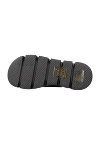 Suela de sandalia de goma negra con un patrón texturizado que presenta surcos distinguidos y un talón circular. Incluye instrucciones de cuidado etiquetadas.