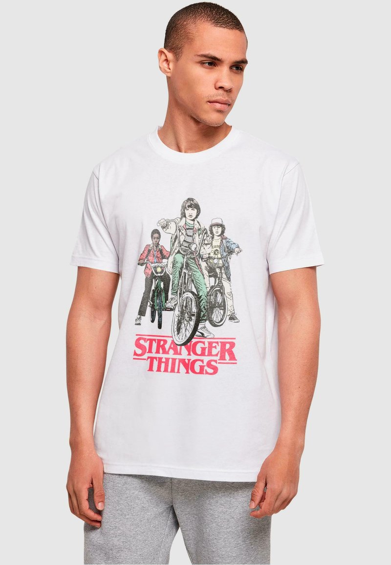 White cotton t-shirt featuring a graphic print of three characters on bicycles with "STRANGER THINGS" in bold red lettering below.