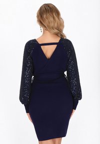 Navyblaues Kleid aus geripptem Stoff mit langen, paillettenbesetzten Puffärmeln und einem Cut-out-Detail am Rücken, abgerundet mit einem Bindegürtel in der Taille.