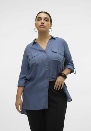 Blouse bleu à boutons avec deux poches poitrine, manches retroussées et ourlet arrondi, portée avec un pantalon noir. Texture de tissu lisse.