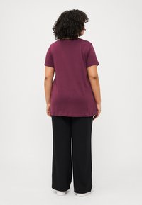 En person med krøllete hår har på seg en maroon t-skjorte og svarte vide bukser, snudd bort fra en ensfarget hvit bakgrunn.
