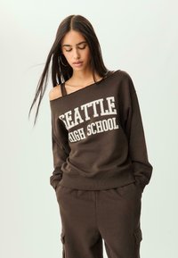Felpa marrone con "SEATTLE HIGH SCHOOL" in testo bianco; presenta un design scollato e polsini a costine. Abbinata a pantaloni cargo coordinati.