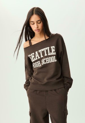 Bruine sweatshirt met "SEATTLE HIGH SCHOOL" in witte tekst; heeft een off-shoulder ontwerp en geribbelde cuffs. Gekleed met bijpassende cargobroek.