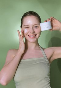 Mieto puhdistusbalmi valkoisessa, pyöreässä rasiassa, jota pidetään henkilön vieressä, joka levittää voidetta kasvoilleen. Taustalla on yhtenäinen vihreä väri.