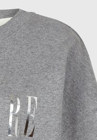 Sudadera gris con una tela texturizada, que presenta una impresión metálica plateada reflectante de las letras "RE" en la parte frontal.